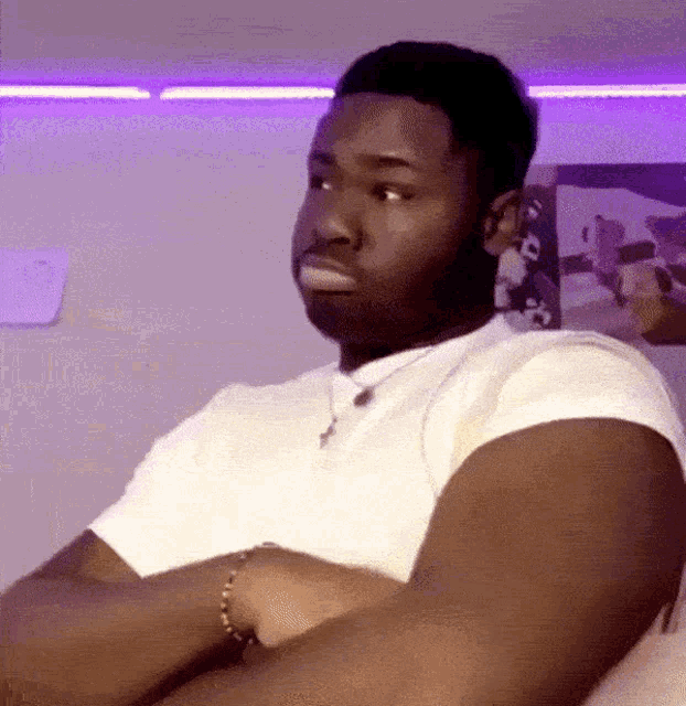 a man in a white shirt is sitting on a bed with his arms crossed and making a funny face .