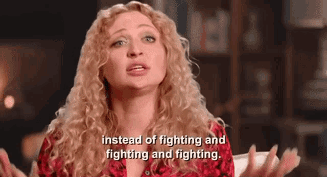 a woman with curly hair is sitting in front of a bookshelf and talking about fighting and fighting .