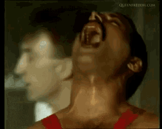 a man in a red tank top is screaming with his mouth open in front of another man .