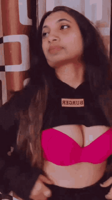 a woman wearing a pink bra and a black hoodie is standing in front of a curtain and looking at the camera .
