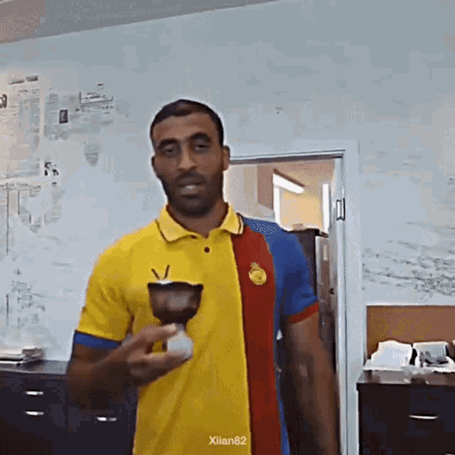 a man in a yellow blue and red shirt is holding a cup with the name xilian82 on the bottom right