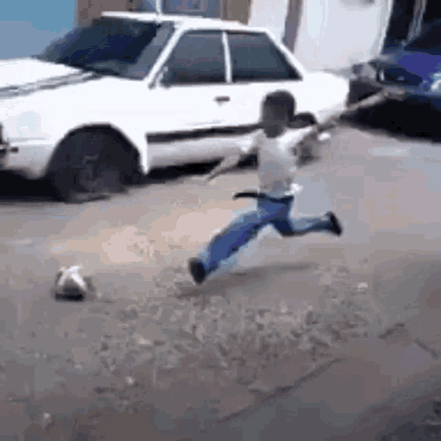 a young boy is kicking a soccer ball on a sidewalk in front of a white car .
