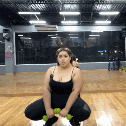 a woman squatting down with dumbbells in front of a sign that says muse zone