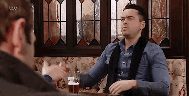 two men are sitting at a table in front of a stained glass window with itv written on the bottom right corner .