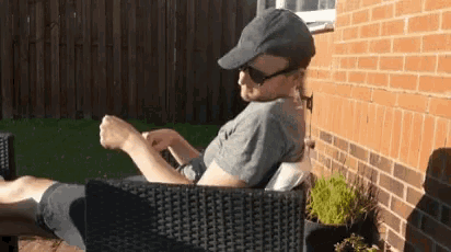 a man wearing sunglasses and a hat is sitting in a chair in front of a brick building .