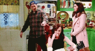 a man in a plaid shirt is pointing at a woman sitting at a table in a kitchen .