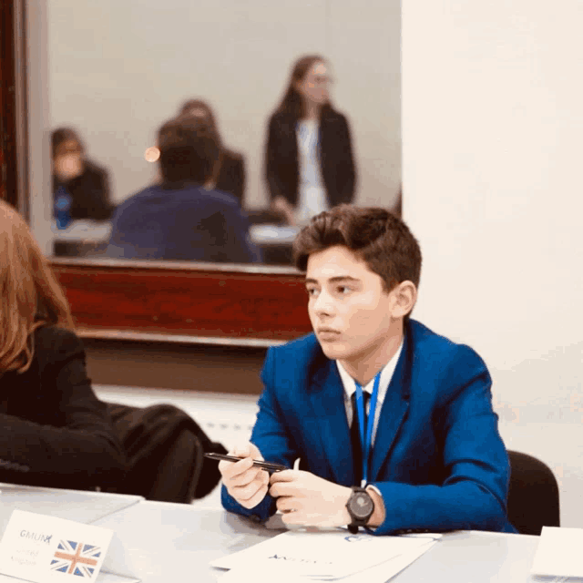 a young man in a blue suit sits at a table with a sign that says gamak on it