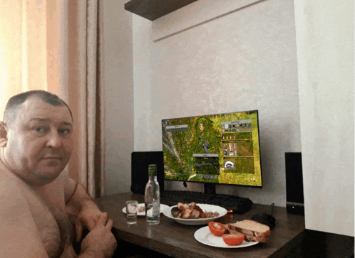 a shirtless man sits at a table with a computer monitor and food