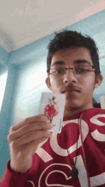 a young man wearing glasses and a red supreme sweatshirt holds up a playing card