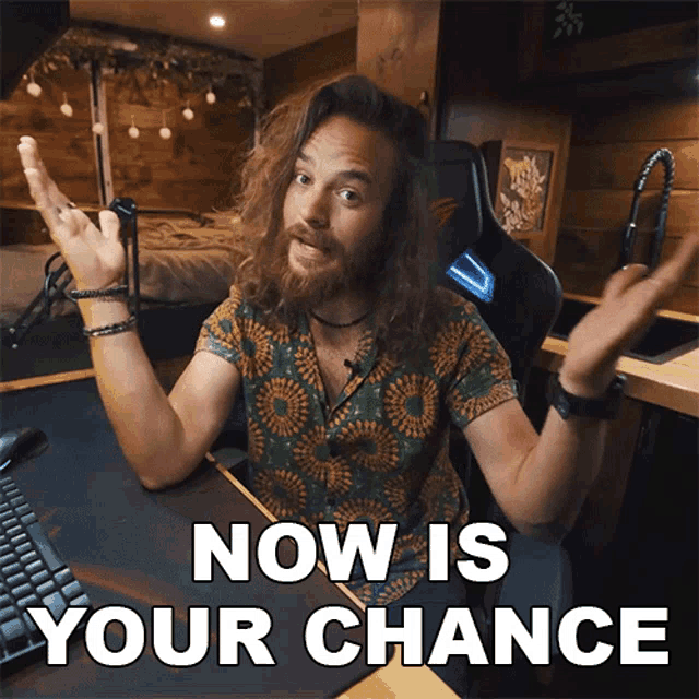a man with long hair and a beard sitting at a desk with the words now is your chance above him