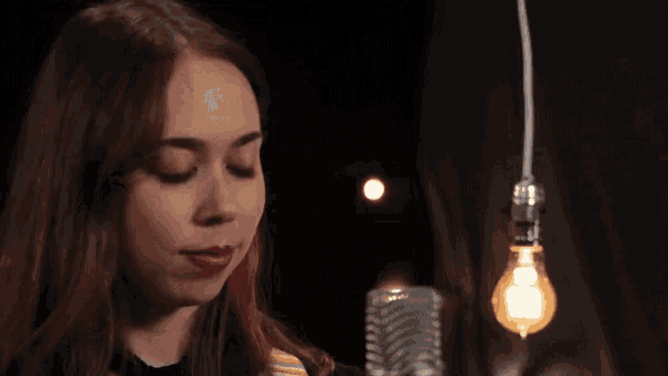 a woman singing into a microphone with a light bulb hanging from the ceiling behind her