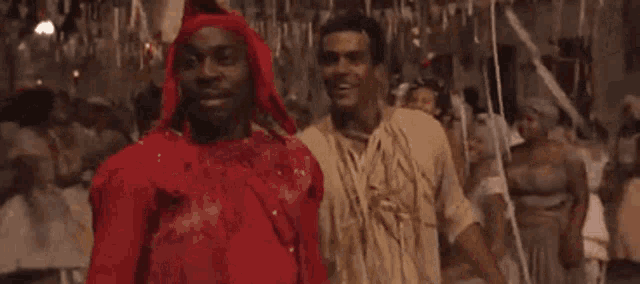 two men are standing next to each other in a crowded room . one of the men is wearing a red robe .