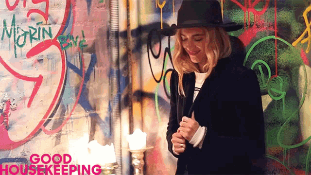 a woman in a black hat stands in front of a wall with graffiti on it that says " good housekeeping "