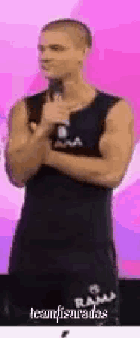 a man in a black tank top is standing in front of a pink background with his hands folded .
