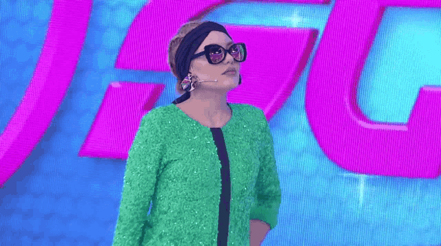 a woman in a green dress and sunglasses stands in front of a purple and blue background with the letter g