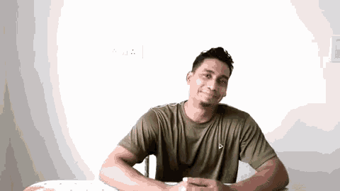 a man in a green t-shirt is sitting at a table with his hands folded and smiling .