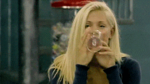 a blonde woman is drinking from a glass in front of a gumball machine .