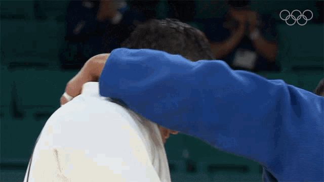 a man in a blue jacket is hugging another man with the olympic rings visible in the background