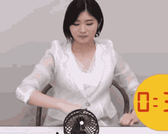 a woman in a white shirt is sitting at a table with a small fan