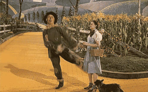 a woman in a blue dress stands next to a scarecrow on a yellow brick road