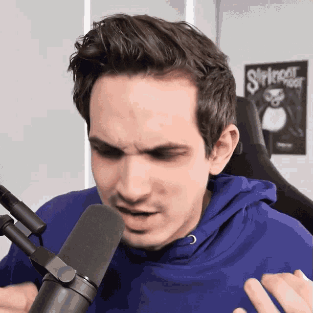 a man wearing a blue hoodie is talking into a microphone with a stringer poster behind him