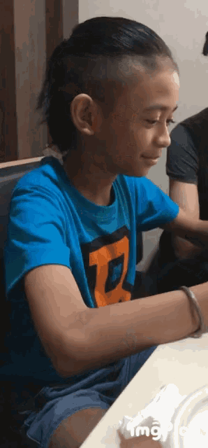 a boy wearing a blue shirt with the letter d on it sits at a table