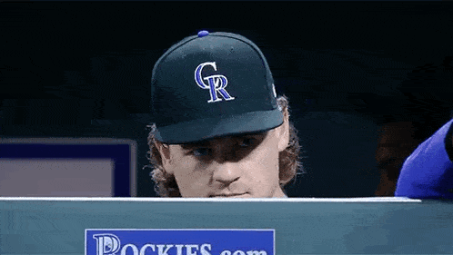a man wearing a rockies baseball cap is looking at a laptop .