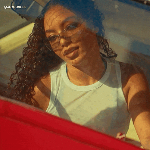 a woman wearing glasses and a white tank top is looking out the window of a red car