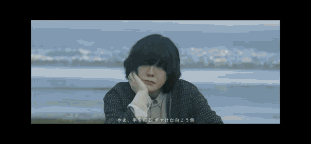 a man with short black hair is sitting on the beach with his hand on his chin .