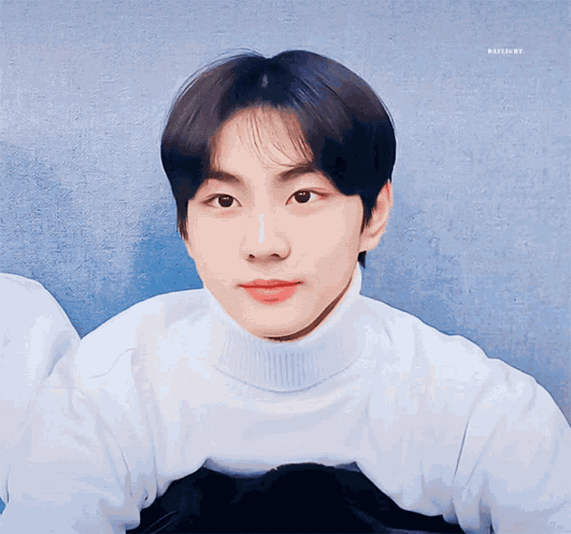 a young man wearing a white turtleneck sweater looks at the camera with a blue background