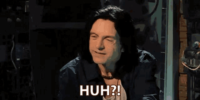 a man with long hair is sitting in a dark room with his eyes closed and says huh .
