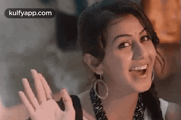 a woman is smiling and waving her hand while wearing a necklace and earrings .