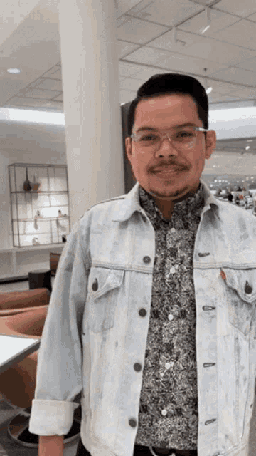 a man wearing glasses and a denim jacket is smiling