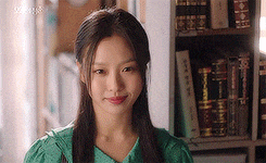 a young woman in a green dress is standing in front of a bookshelf .