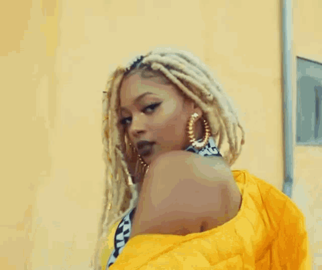 a woman with blonde dreadlocks and hoop earrings is wearing a yellow jacket and earrings .
