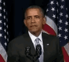 barack obama is giving a speech in front of an american flag while standing in front of a microphone .