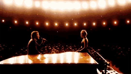 a man singing into a microphone while a woman sits at a piano in front of a crowd