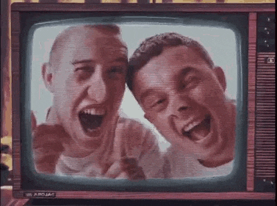 two men are laughing and pointing at the camera in front of a television screen .