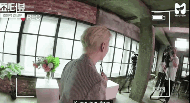 a man with blonde hair is standing in front of a window while a cameraman takes a picture of him