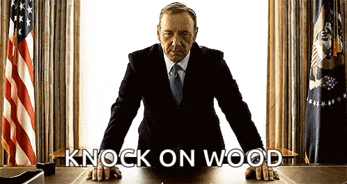a man in a suit and tie is leaning on a desk with the words " knock on wood " written on it .