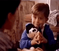 a young boy is sitting at a table eating a hamburger with a stuffed mickey mouse .