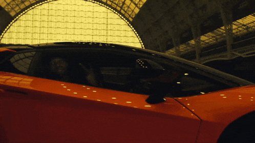 a man sits in an orange sports car in front of a large building