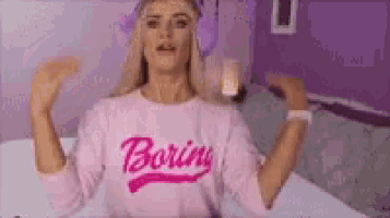 a woman is sitting on a bed wearing a pink shirt that says boring .