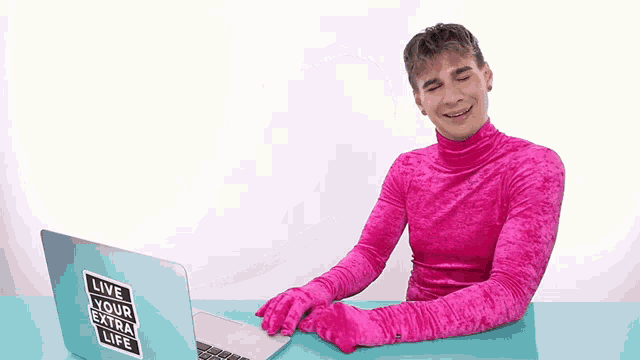 a man in a pink turtleneck is sitting in front of a laptop with a sticker that says live your extra life