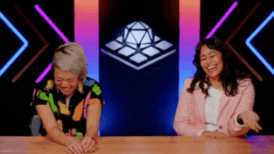 two women are sitting at a table laughing in front of a screen with a cube on it .