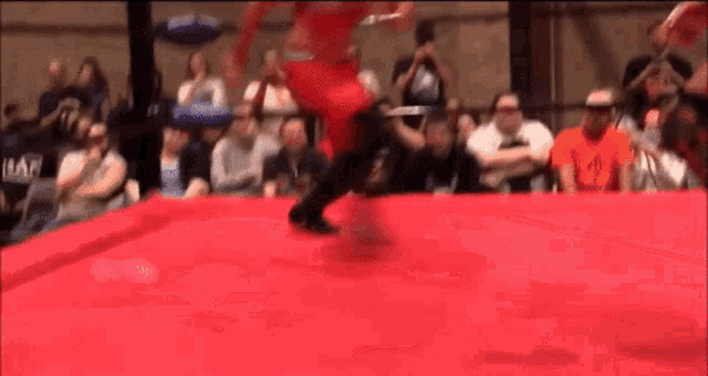 a wrestler is jumping over a red mat in a ring while a crowd watches .