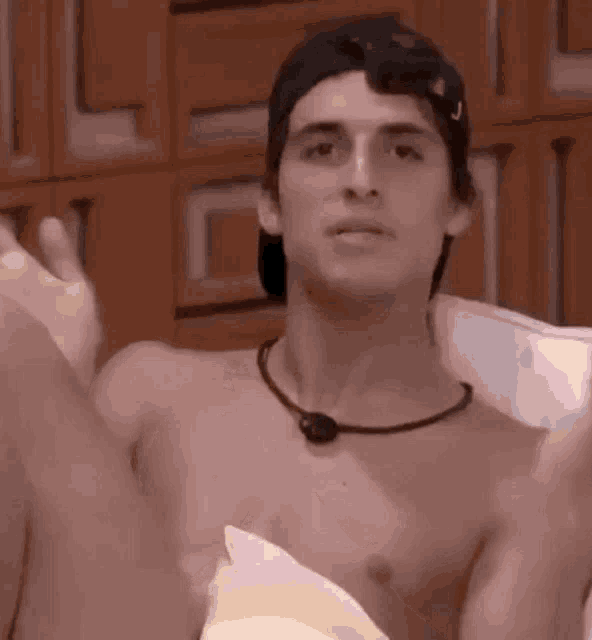 a shirtless man wearing a hat and a necklace is sitting on a bed with his arms outstretched .
