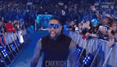 a man wearing sunglasses and a black vest is standing in front of a crowd of people .