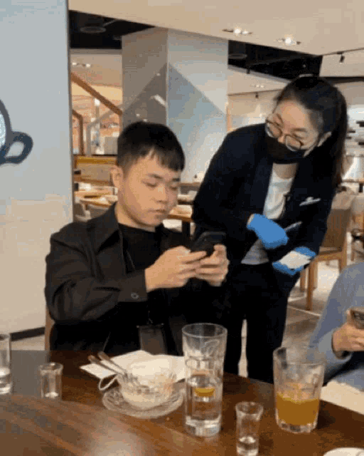a man sits at a table looking at his phone while a woman stands behind him