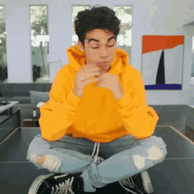 a young man in a yellow hoodie is sitting on a table with his legs crossed and eating something .